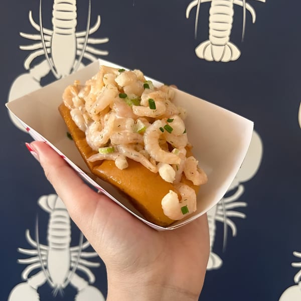 Hand holding a shrimp roll in a paper tray against a blue background with white lobster patterns.