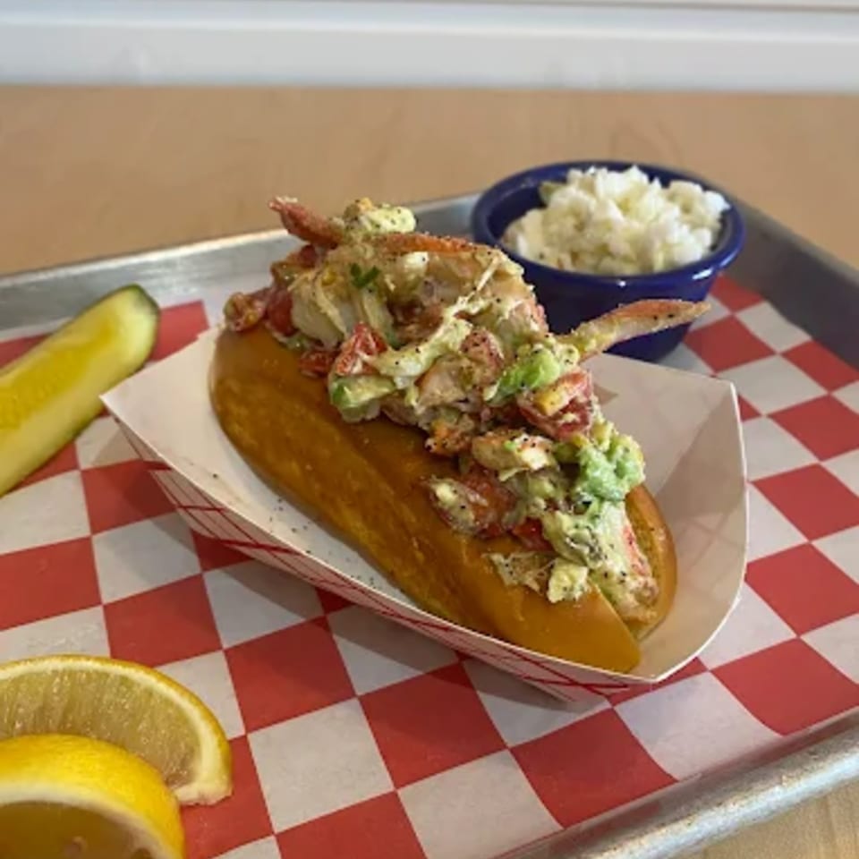 A Boca Lobster Roll with avocado, tomato, and coleslaw, served with a pickle and lemon slices.