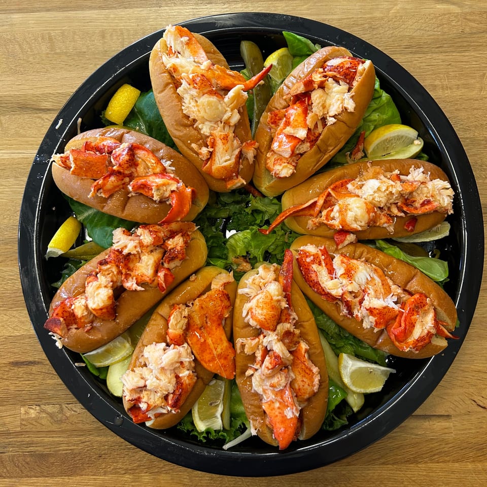 Eight Boca Lobster Roll Company lobster rolls arranged on a black platter with lettuce and lemon wedges.