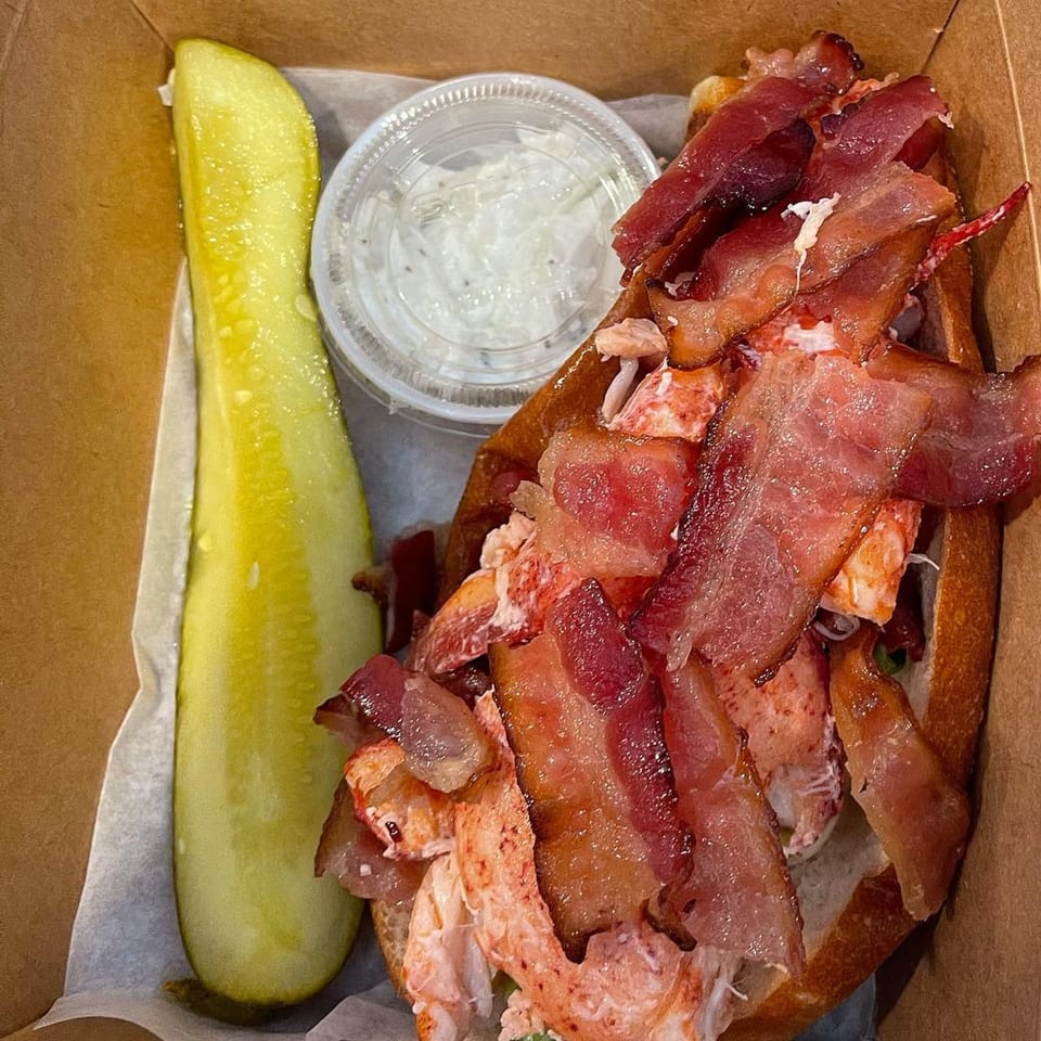 A Boca Lobster Roll with bacon, a pickle spear, and a side of sauce in a takeout container.