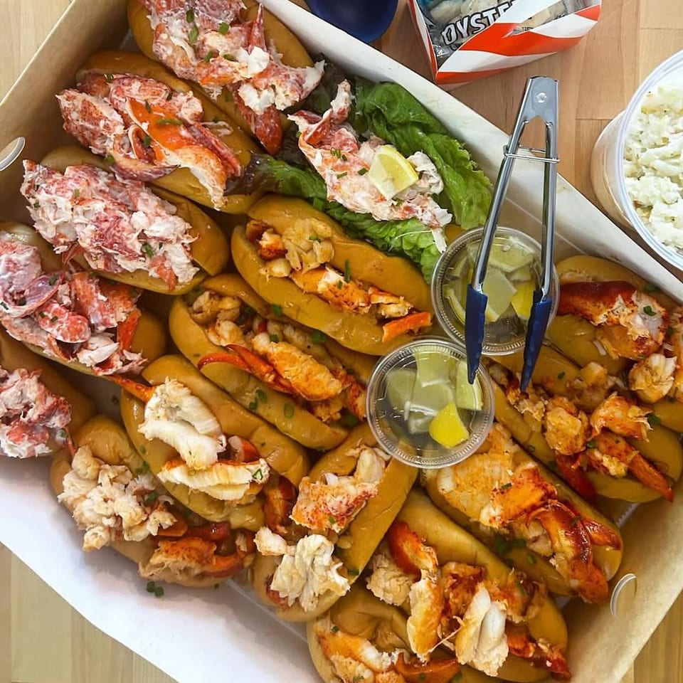 A catering box filled with numerous Boca Lobster Roll Company lobster rolls, some with lettuce and lemon.