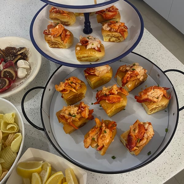 A two-tiered tray of mini lobster rolls, with chips, lemon wedges, and desserts on a counter.